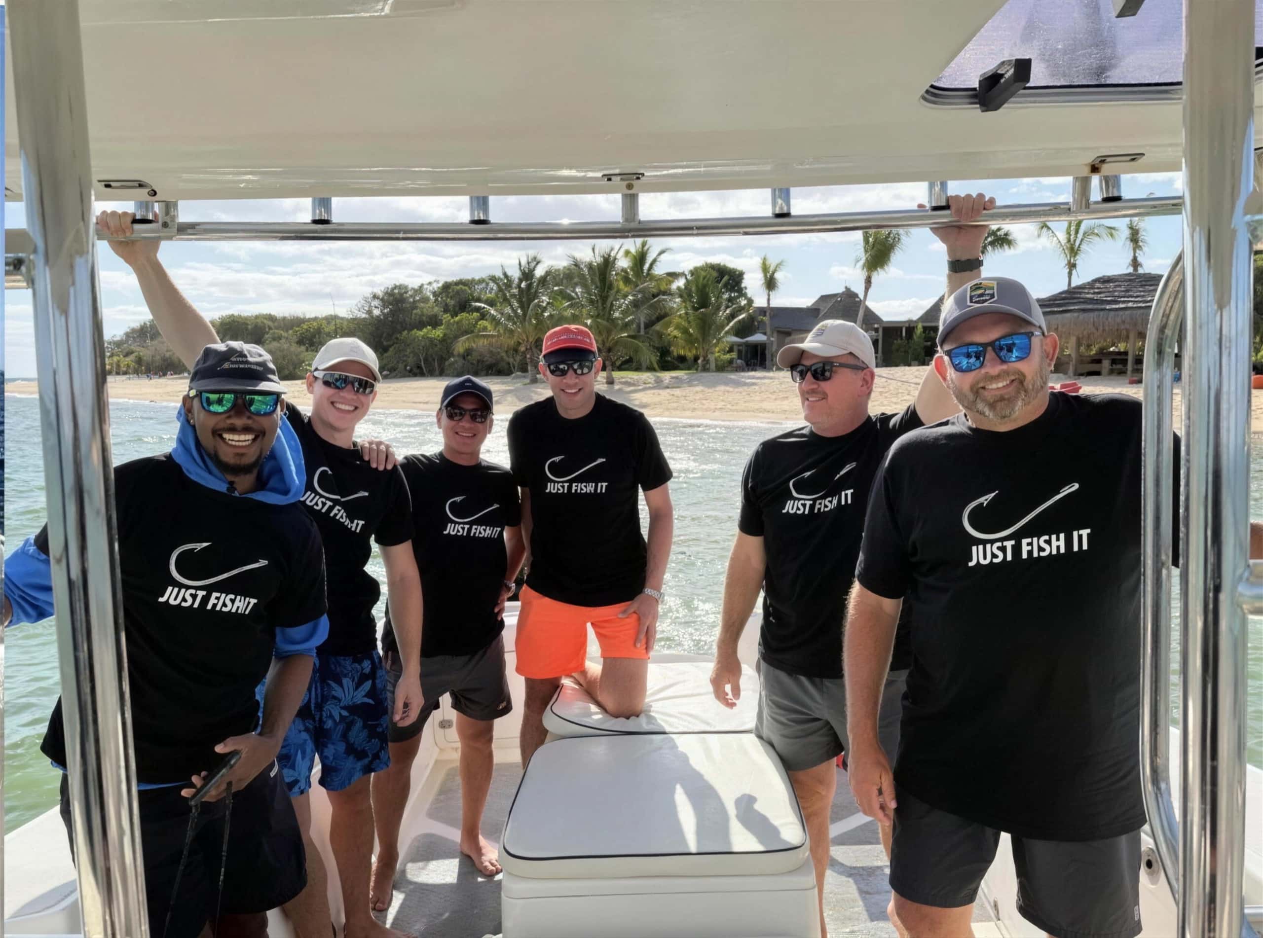 Six guests in matching fishing shirts on the deck of a boat, moored off a beach