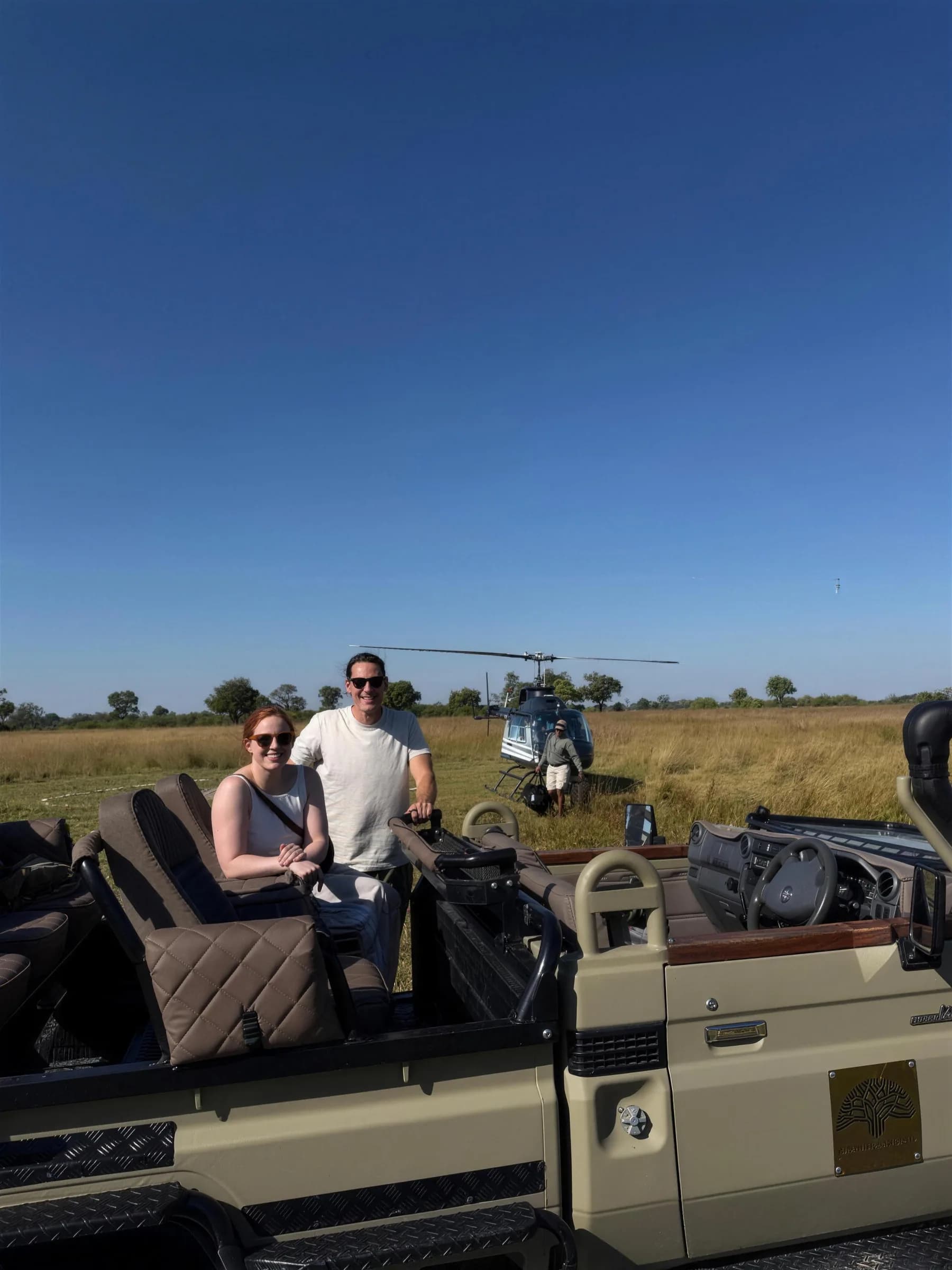 David and Sam Schupp in an open safari vehicle in the Okavango Delta, a helicopter waiting in the grass behind them